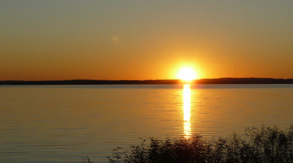 Loppukesän auringon lasku. Taivas ja järvimaisema hehkuu oranssin sävyissä.
