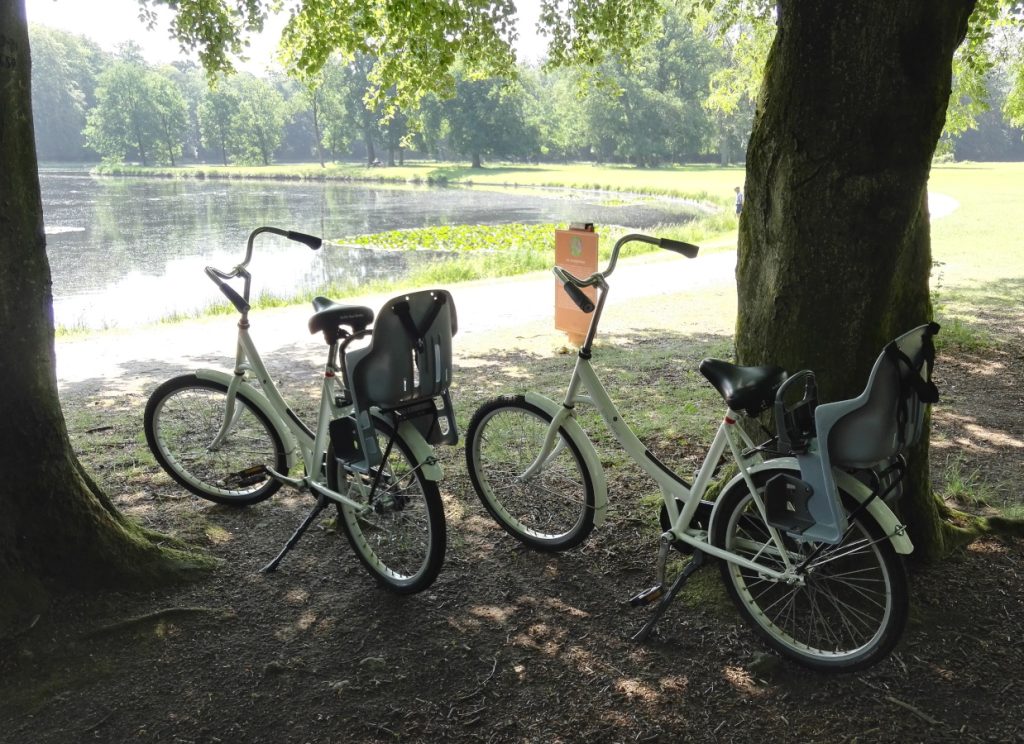 Valkoiset polkupyörät puiden välissä. Edessä näkyy hiekkapolku ja lampi.