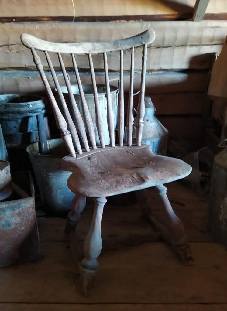 Lähikuva puisesta vanhasta tuolista. Takana metalliastioita ja hirsiseinä.